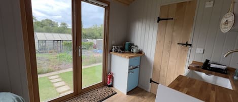 Une cuisine compacte dotée d’une porte-fenêtre coulissante donnant sur un jardin, d’un petit réfrigérateur et d’un plan de travail en bois.