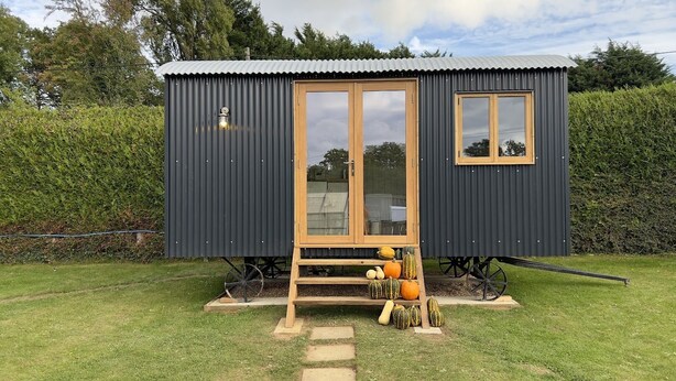 A small, dark-colored metal structure with a wooden door and window, a step leading up to it, and a wheelbarrow with pumpkins and gourds in front. - Luxury Shepherds Hut, W/large Garden, Firepit, & Wi-fi @ Cedar Gables Campsite (Flimwell, Wadhurst)
