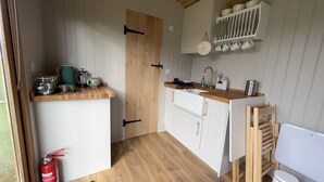 A compact kitchen with white cabinets, a wooden countertop, and a white sink. There is a fire extinguisher on the floor, and a shelf with dishes above the countertop. - Luxury Shepherds Hut, W/large Garden, Firepit, & Wi-fi @ Cedar Gables Campsite (Flimwell, Wadhurst)