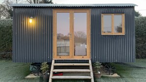 A small, dark-colored metal shed with a wooden door and window, a small set of steps leading up to it, and two potted plants on either side. - Luxury Shepherds Hut, W/large Garden, Firepit, & Wi-fi @ Cedar Gables Campsite (Flimwell, Wadhurst)