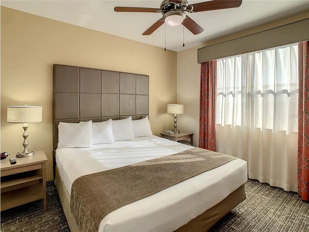 A hotel room with a large bed, two bedside tables, a ceiling fan, and a window with curtains.