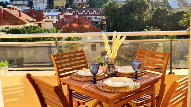 Ein Holztisch ist gedeckt mit Tellern, Gläsern und einer Vase mit getrockneten Pflanzen auf einem Balkon mit Blick auf Gebäude.