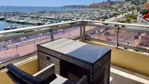 Un balcón con mobiliario de mimbre, una mesa de vidrio y vistas a un puerto deportivo con barcos y un paisaje urbano al fondo.