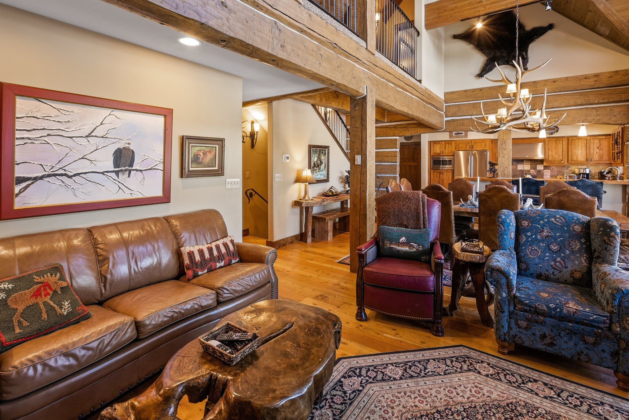 A spacious living room with a leather couch, patterned armchairs, and a wooden coffee table. A staircase leads to an upper level, and the room features wooden beams and a chandelier.