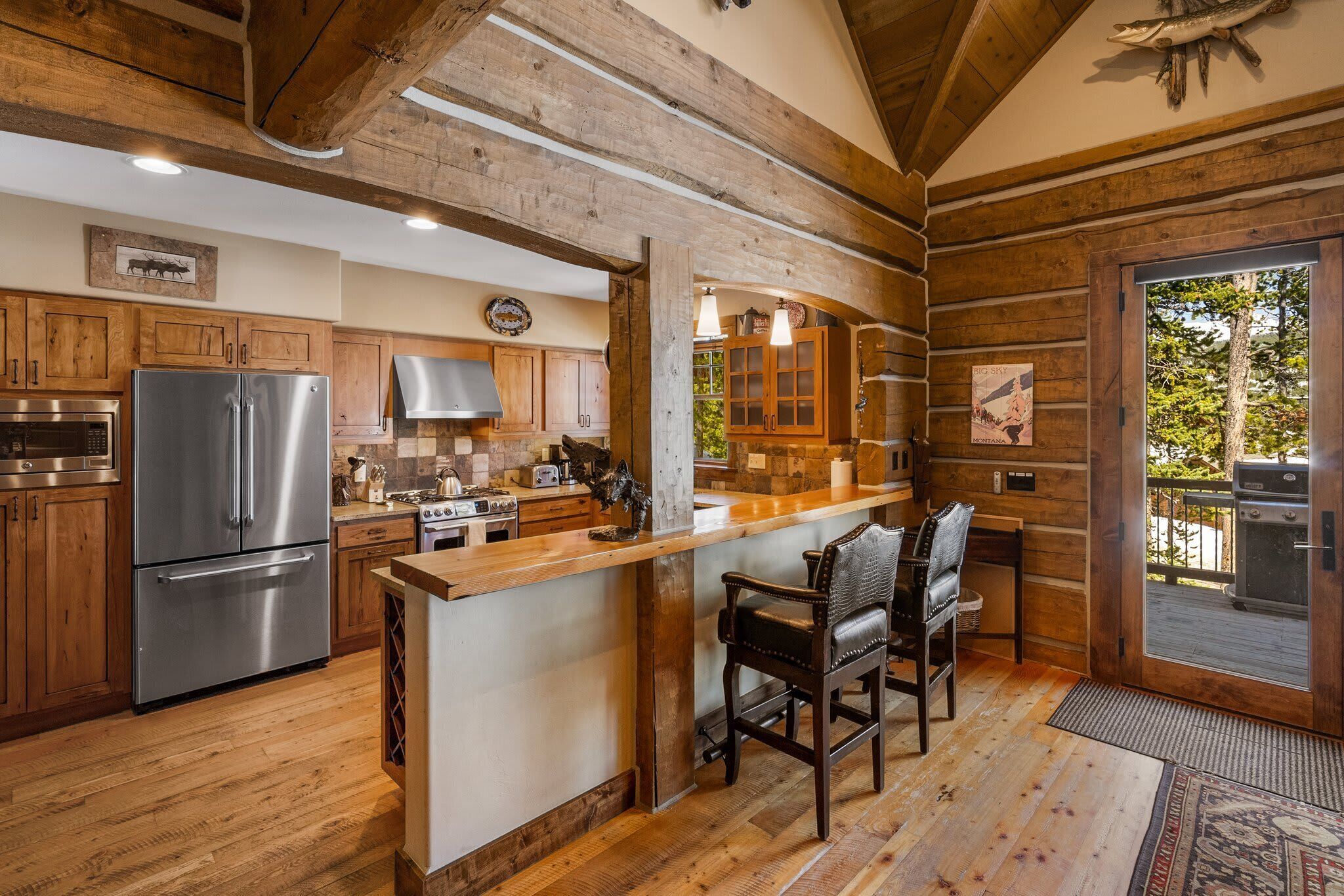 A rustic kitchen with wooden beams, a stainless steel refrigerator, wooden cabinets, a bar area with chairs, and a view of a grill outside.