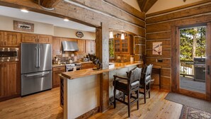 A rustic kitchen with wooden beams, a stainless steel refrigerator, wooden cabinets, a bar area with chairs, and a view of a grill outside. - Luxury Powder Ridge Cabin - Ski-In/Ski-Out Access (Big Sky)