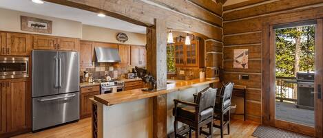 A rustic kitchen with wooden beams, a stainless steel refrigerator, wooden cabinets, a bar area with chairs, and a view of a grill outside.