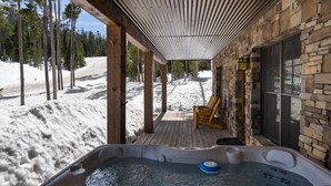 A hot tub with a wooden deck and stone walls, overlooking a snowy landscape with trees. - Luxury Powder Ridge Cabin - Ski-In/Ski-Out Access (Big Sky)