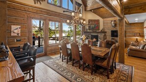 A spacious dining area with a large wooden table and leather chairs, a fireplace, and a view of the outdoors through large windows. - Luxury Powder Ridge Cabin - Ski-In/Ski-Out Access (Big Sky)