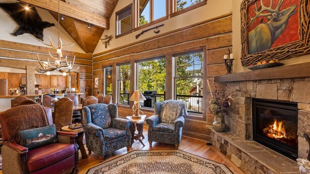 A spacious living room with a stone fireplace, large windows, and rustic decor. - Luxury Powder Ridge Cabin - Ski-In/Ski-Out Access (Big Sky)