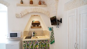 A compact kitchen with a microwave, sink, and a small refrigerator. There is a shelf above the sink with decorative items and a small window above the microwave.
