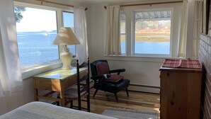 A room with a bed, a desk with a lamp, a chair, and a view of the water. - Cozy Waterfront Cottage, Henry Cove. Winter Harbor (Winter Harbor)