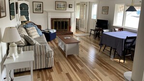 A spacious living room with a fireplace, a dining area with a table and chairs, and a seating area with a sofa and a lamp. - Cozy Waterfront Cottage, Henry Cove. Winter Harbor (Winter Harbor)