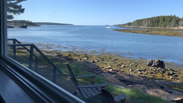 A wooden deck with a chair and a table, overlooking a calm body of water with a distant shoreline and trees. - Cozy Waterfront Cottage, Henry Cove. Winter Harbor (Winter Harbor)