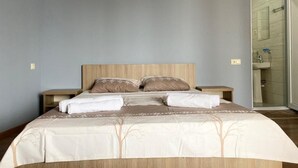 A neatly made bed with a wooden headboard, two bedside tables, and a bathroom visible through an open door. - Hotel Charli (Kobuleti)