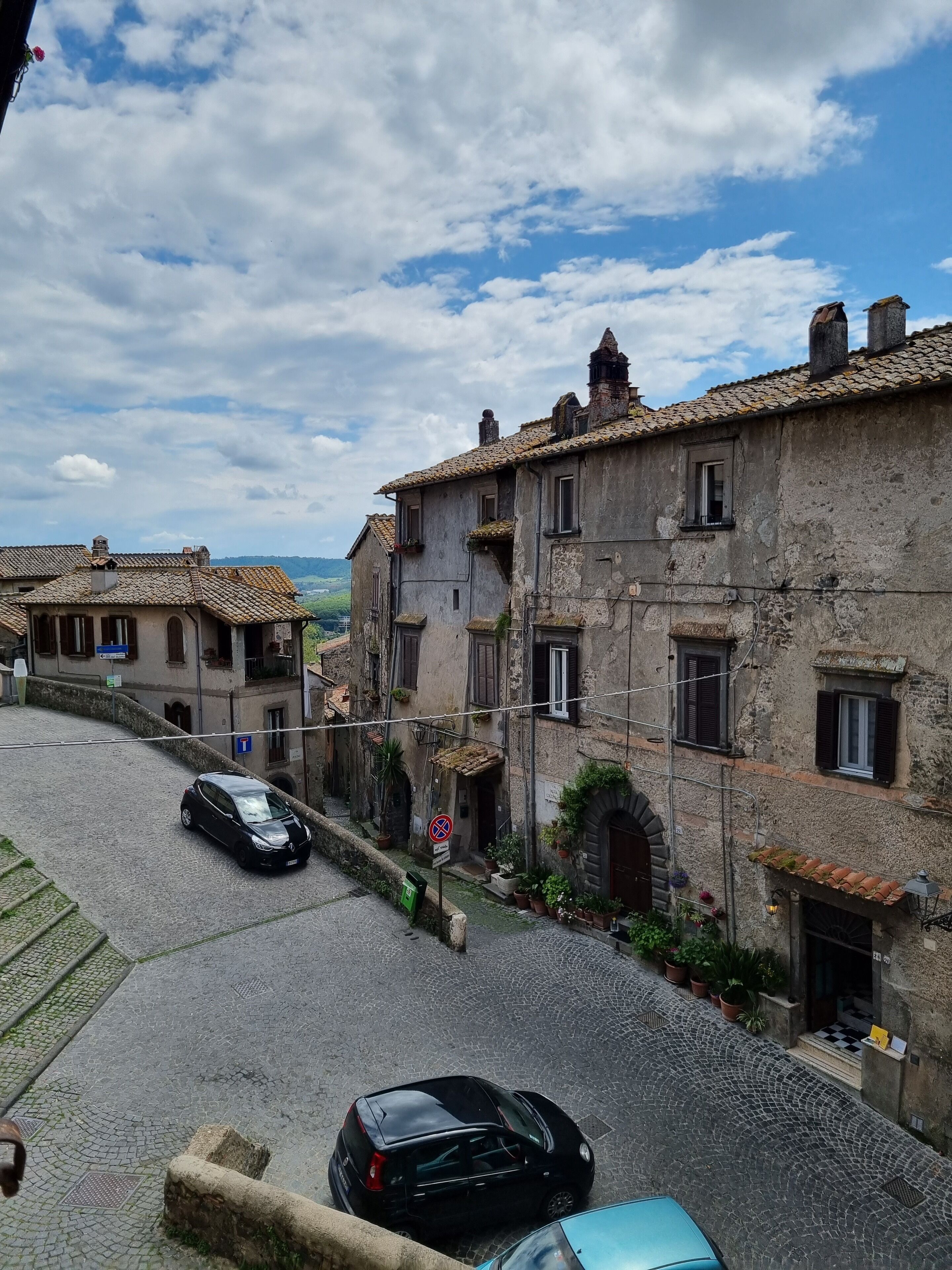 A narrow cobblestone street lined with historic stone buildings, a black car parked on the side, and a blue car in the foreground.