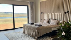 A modern bedroom with a large bed, a view of a landscape through a sliding door, and a potted plant on a wooden table. - Spacious villa in Golden Circle with amazing view & hot tub! (Reykholt)