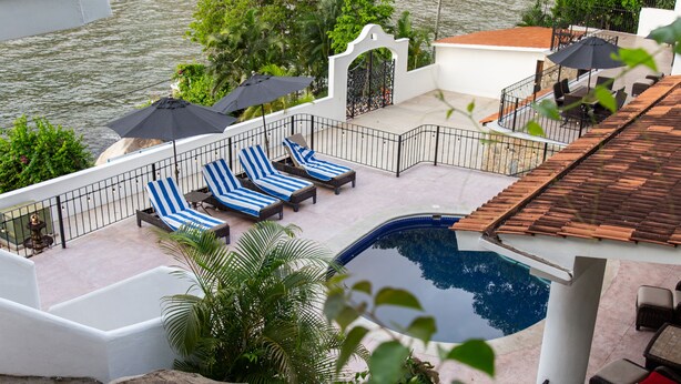 Zona de piscina en la azotea con sillones de descanso y sombrillas, rodeada de vegetación y con vistas a un cuerpo de agua.