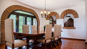 Un comedor con mesa de madera y sillas blancas tapizadas, arcos de ladrillo y un ventilador de techo.
