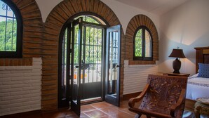 Una habitación con un amplio arco de entrada, una silla de madera, una cama con almohada estampada y una lámpara sobre una mesita de noche.