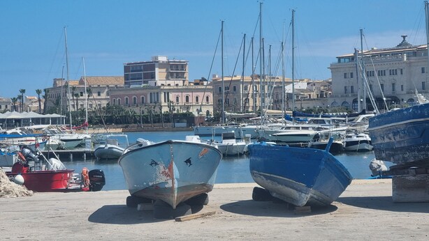 Un puerto deportivo con barcos amarrados, un primer plano de arena y edificios al fondo.