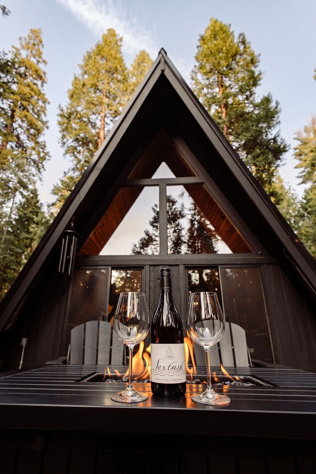 A wooden cabin with a triangular roof, a fireplace, and two wine glasses and a bottle on the hearth.