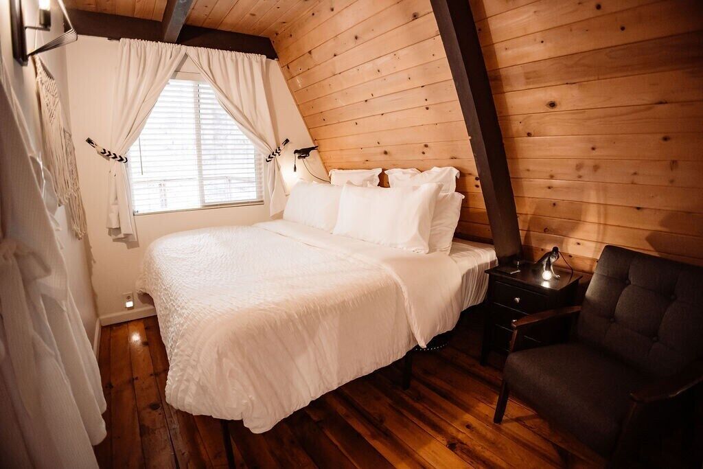 Une chambre confortable avec un plafond en bois, un lit recouvert de linge de lit blanc, une table de chevet avec une lampe et un fauteuil sombre.