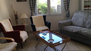 A living room with a red armchair, a white armchair, a grey sofa, a glass coffee table, and a window with checkered curtains. - Maison Très Bien Située,dans Quartier Résidentiel.endroit Très Calme ,près Océan (Biscarrosse plage)