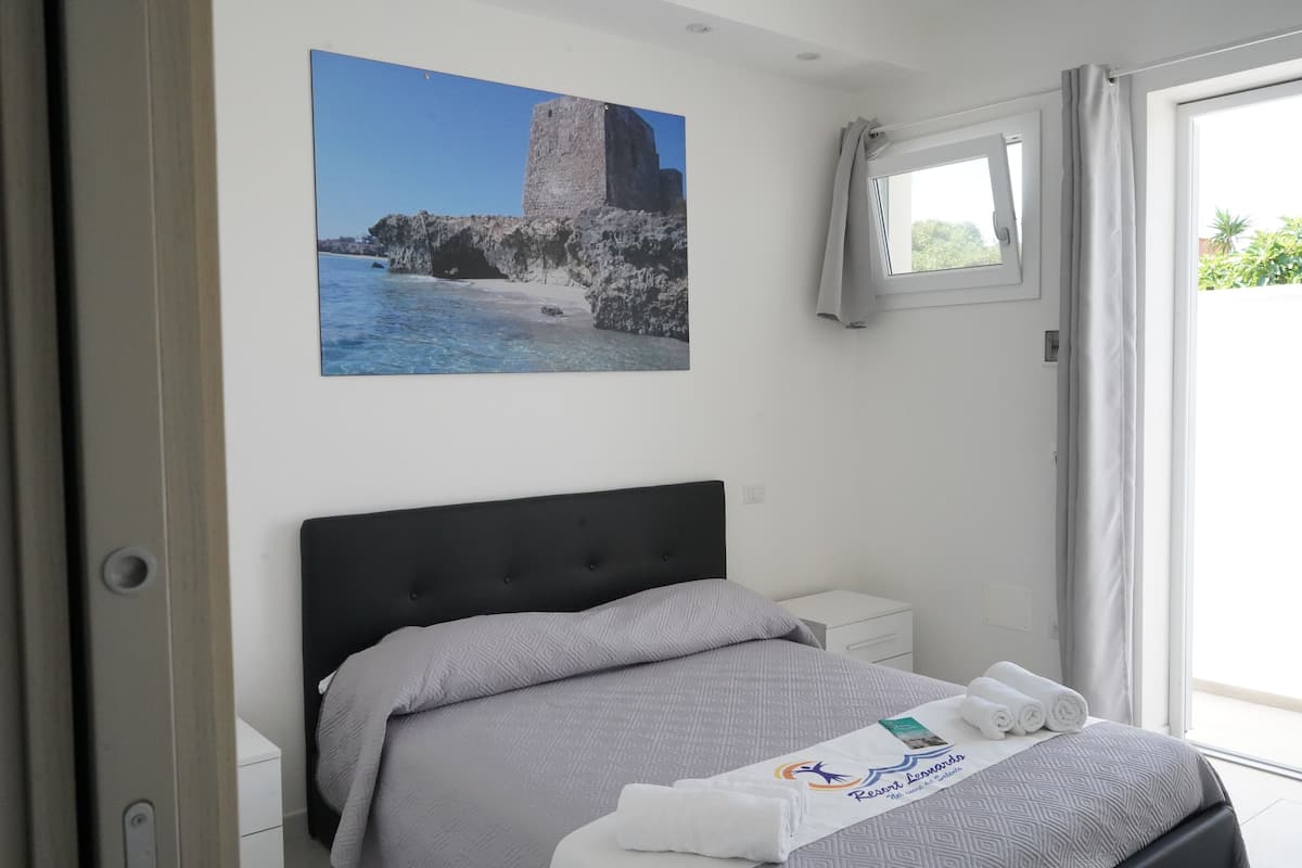 Habitación de hotel con cama, mesita de noche, ventana con cortinas e imagen de escena costera colgada en la pared.