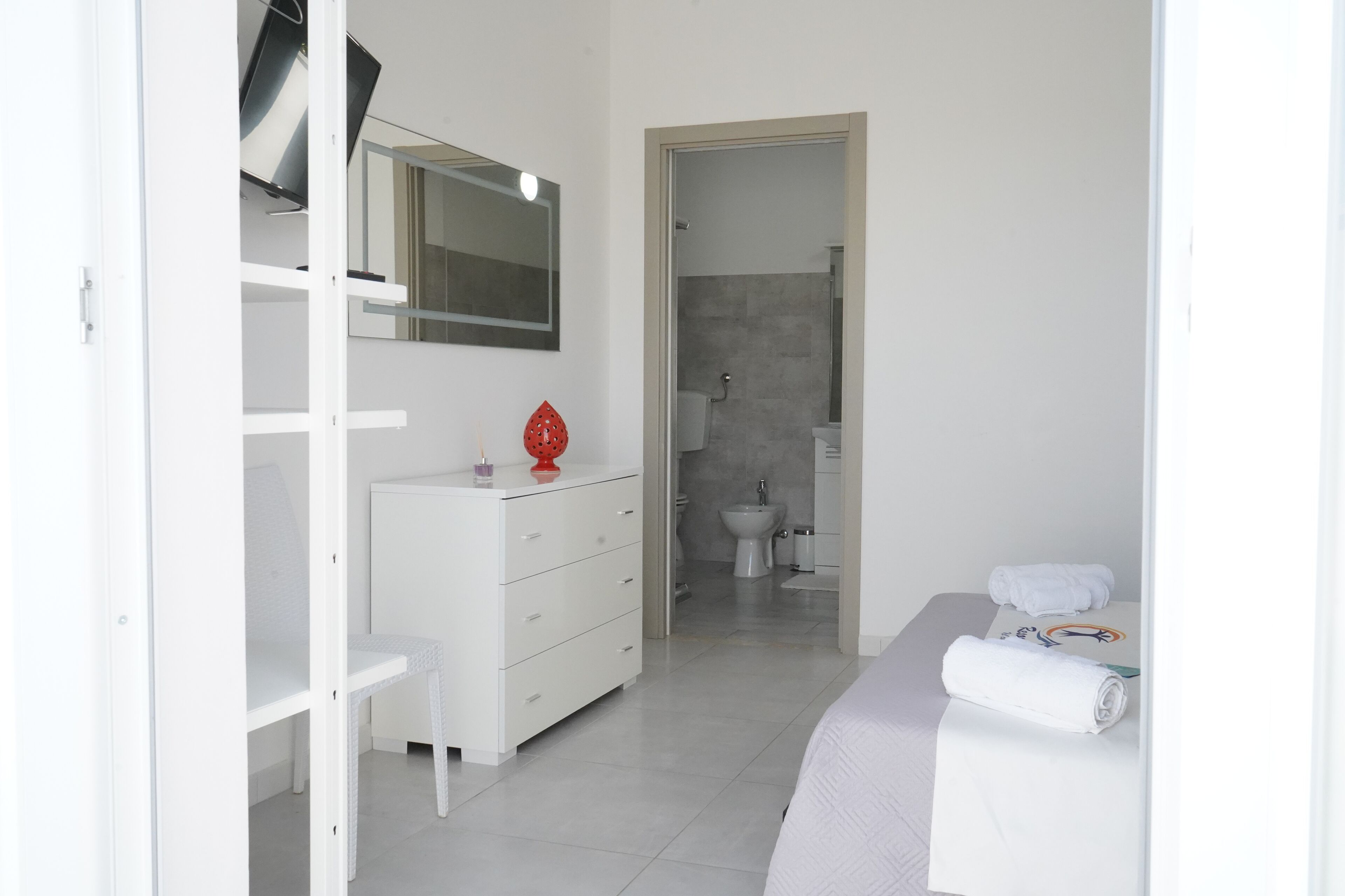 Un dormitorio moderno con cama, cómoda blanca, espejo y baño visible a través de una puerta abierta.