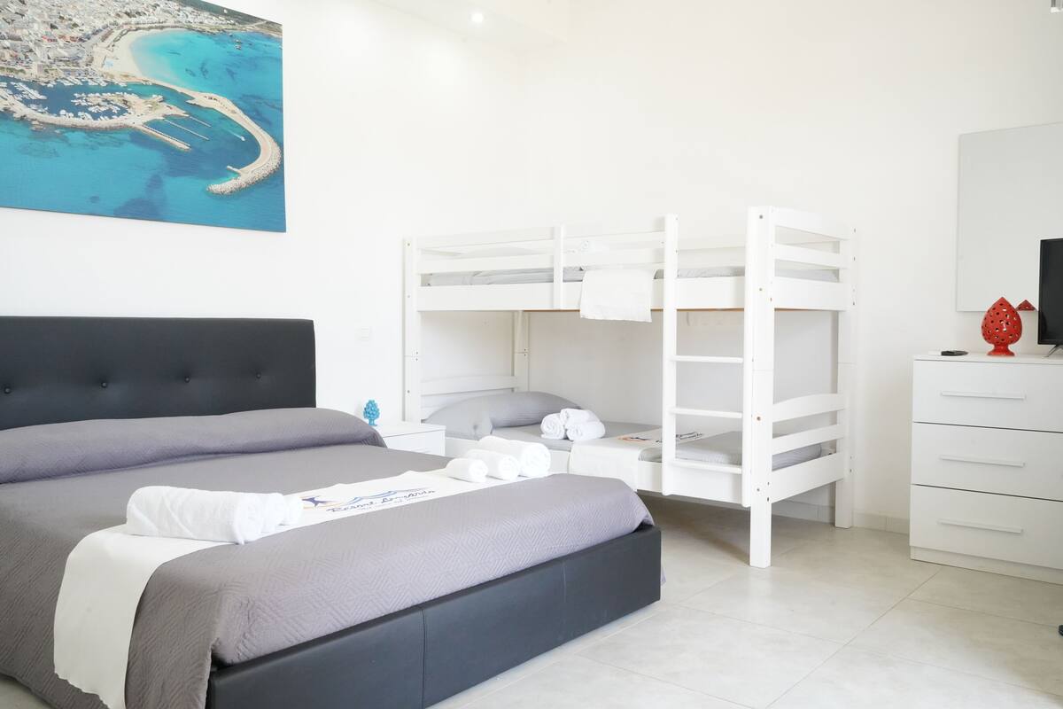 Habitación de hotel con una cama grande, una cama individual superpuesta, un aparador blanco y un cuadro de una escena costera.