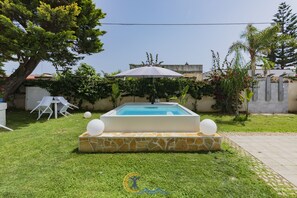 A swimming pool with a stone surround, a white umbrella, and a small garden area with chairs and plants. - Resort Leonardo (Melendugno)