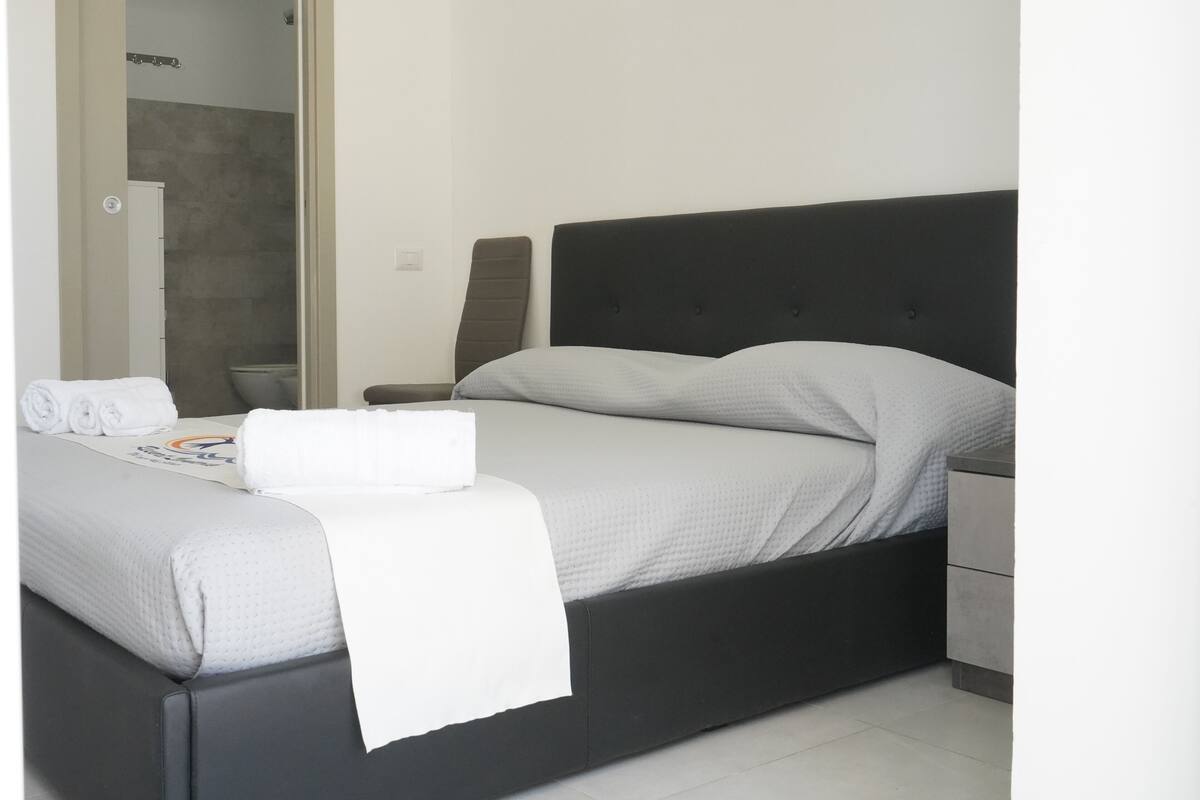 Un dormitorio moderno con cama, mesita de noche y baño a la vista, visible a través de una puerta abierta.