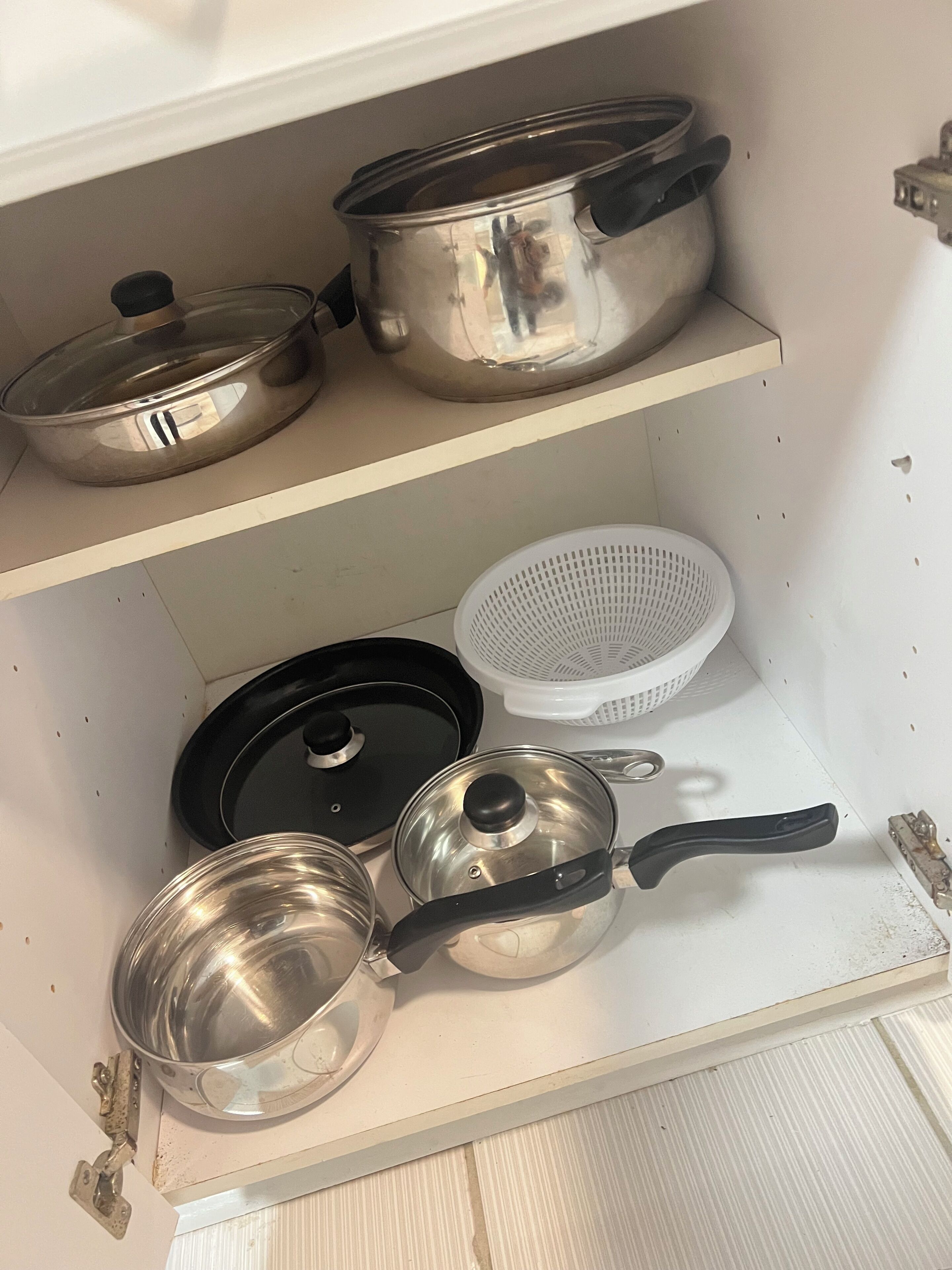 Un armario de cocina con utensilios de acero inoxidable, incluyendo ollas, sartenes y un colador.