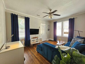 Un salon avec un canapé bleu, un meuble TV blanc, un ventilateur de plafond et une petite table sur laquelle est posée une plante.