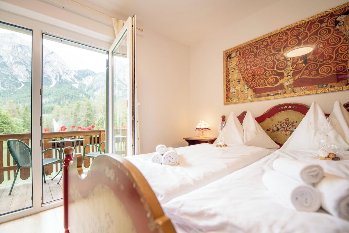 Habitación de hotel con cama, mesita de noche, lámpara y vistas a un paisaje montañoso a través de un gran ventanal.