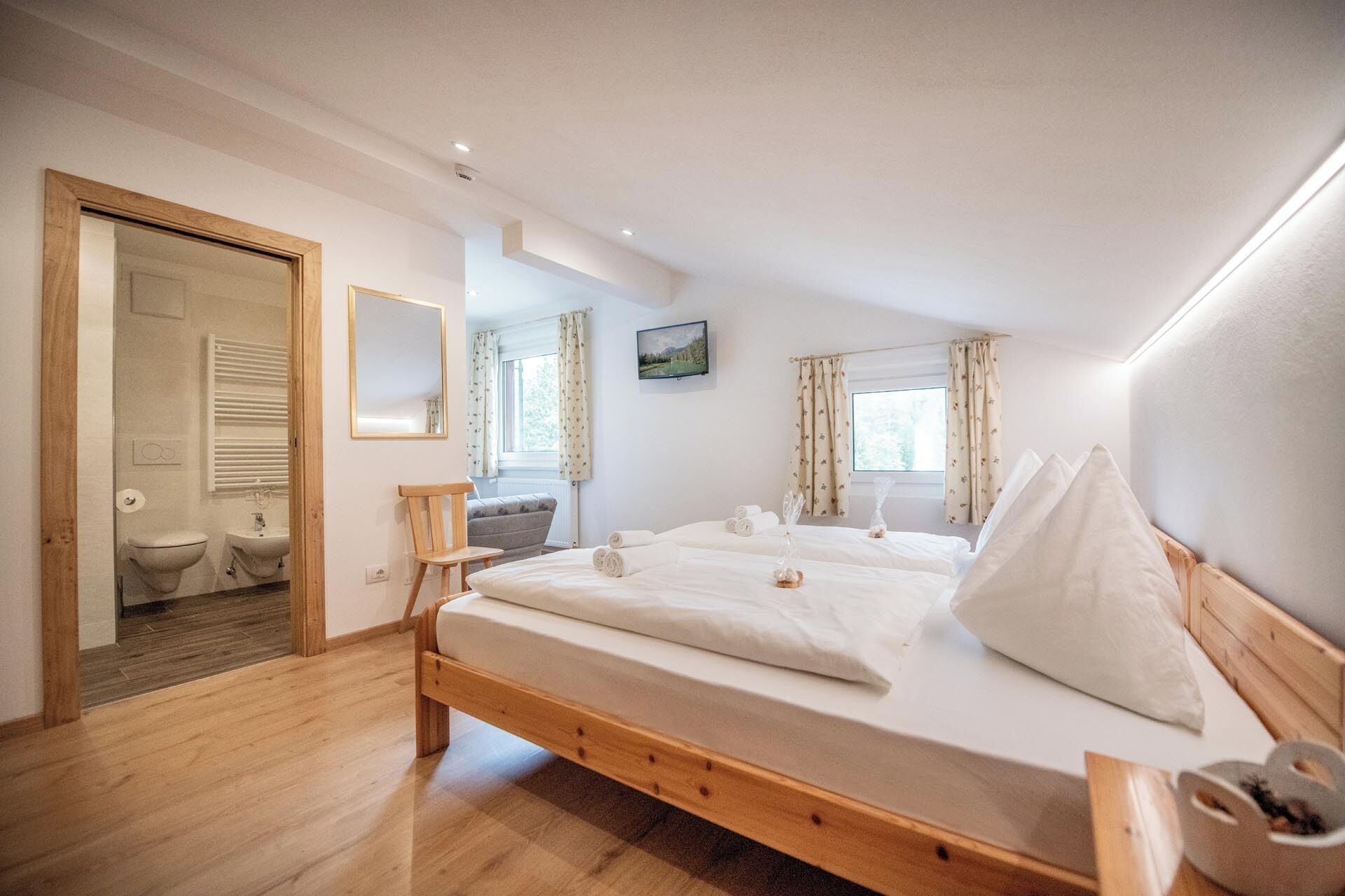 A bedroom with a wooden bed, a small bedside table, a mirror, and a window with curtains.