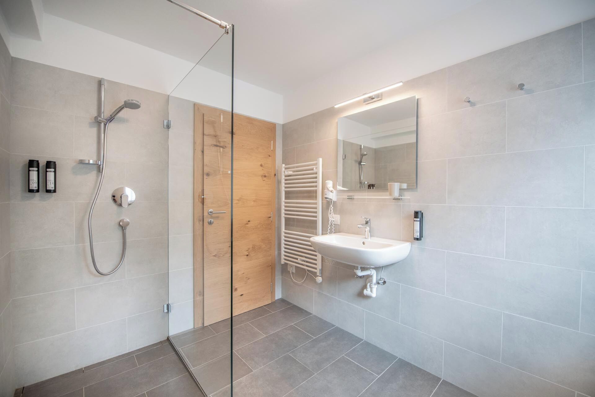 A modern bathroom with a glass shower enclosure, a wooden door, a white sink, and a mirror.