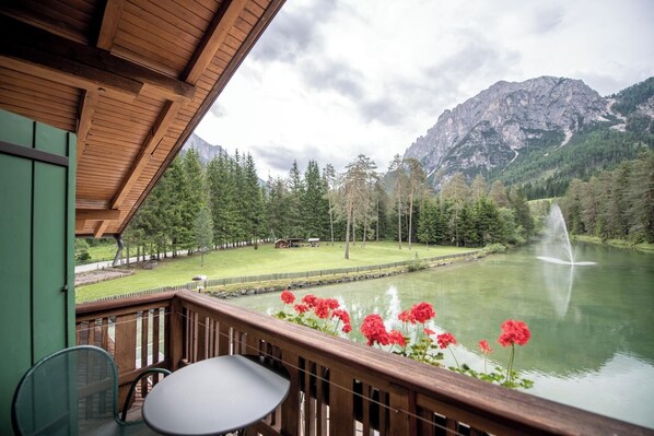 Un balcón de madera con una mesa y sillas da a un lago con una fuente, rodeado de árboles y montañas.