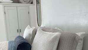 Ein ordentlich bezogenes Bett mit einer gesteppten blauen Decke und mehreren Kissen, ein weißer Kleiderschrank und ein runder Spiegel an der Wand.