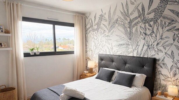 Un dormitorio con una cama, una ventana, una pared con estampado de plantas, una lámpara colgante y una mesita de noche con lámpara.