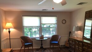 Une pièce comprenant deux fauteuils en osier, une petite table ronde, un meuble en bois et une grande fenêtre avec des stores.