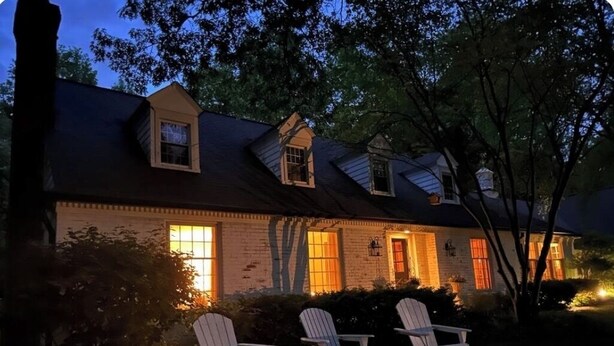 Une maison à deux étages avec une façade en briques, des fenêtres en lucarne et un porche agrémenté de trois fauteuils Adirondack.