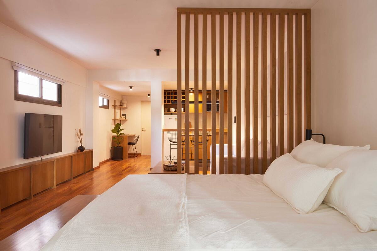 A bedroom with a large bed, wooden slats, a TV, and a view into the kitchen.