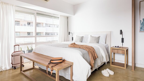 Habitación de hotel con cama, mesita de noche de madera, un banco, maleta y ventana con cortinas.