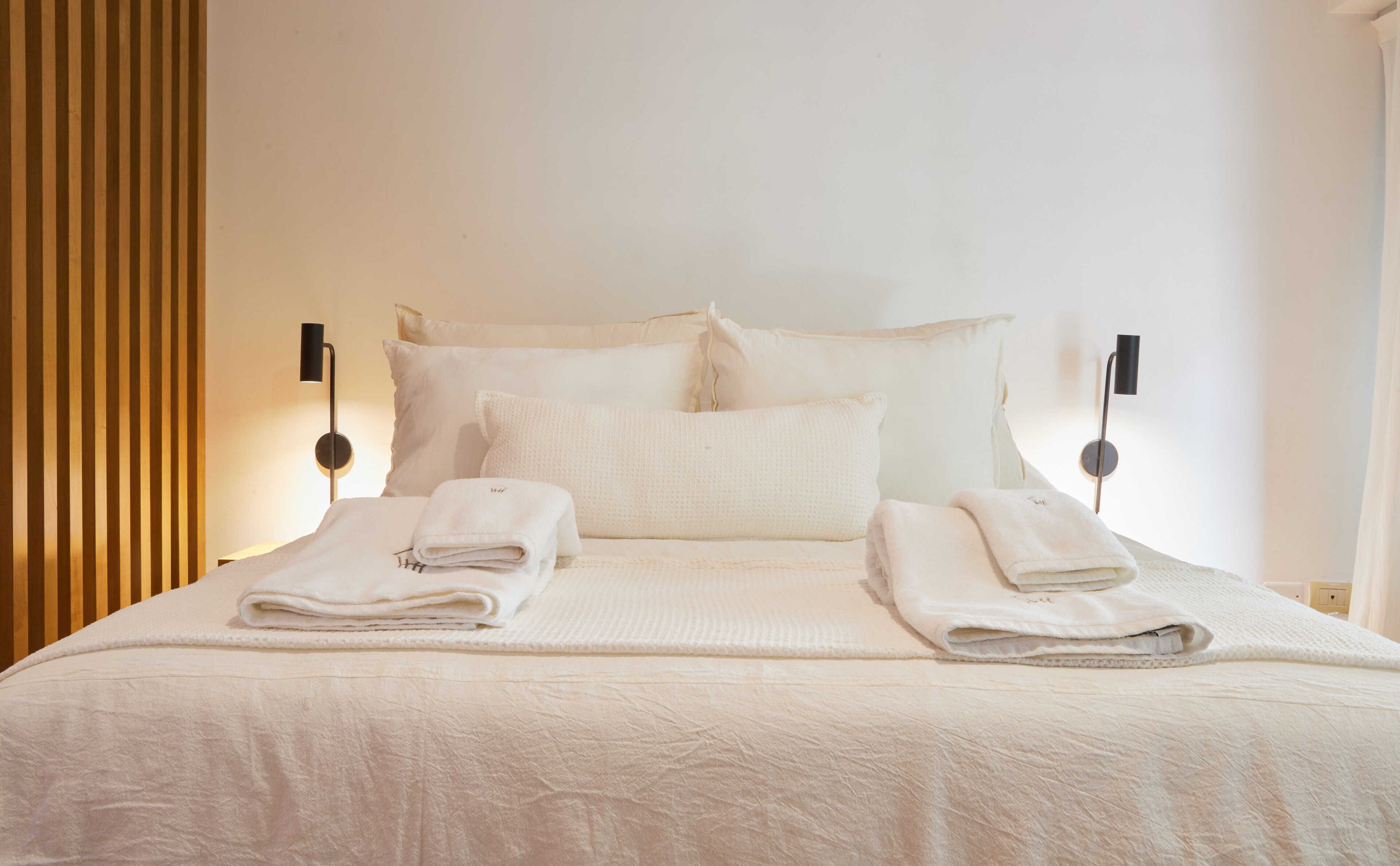 A neatly made bed with white linens and towels, two bedside lamps, and a wooden panel on the left.