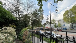 A street view with a black metal fence, trees, and a building in the background. - Studio Face Entrée de Roland Garros (BOULOGNE BILLANCOURT)