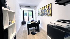 A modern, compact room with a dining table, chairs, a black sofa, a printer, and a large window with a view of the outdoors. - Studio Face Entrée de Roland Garros (BOULOGNE BILLANCOURT)