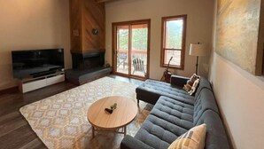 Ein geräumiges Wohnzimmer mit einem großen Sofa, einem Fernseher, einem Kamin und einem großen Fenster.