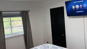 Makuuhuone, jossa on sänky, ikkuna verhoilla, seinälle asennettu televisio ja ovi.